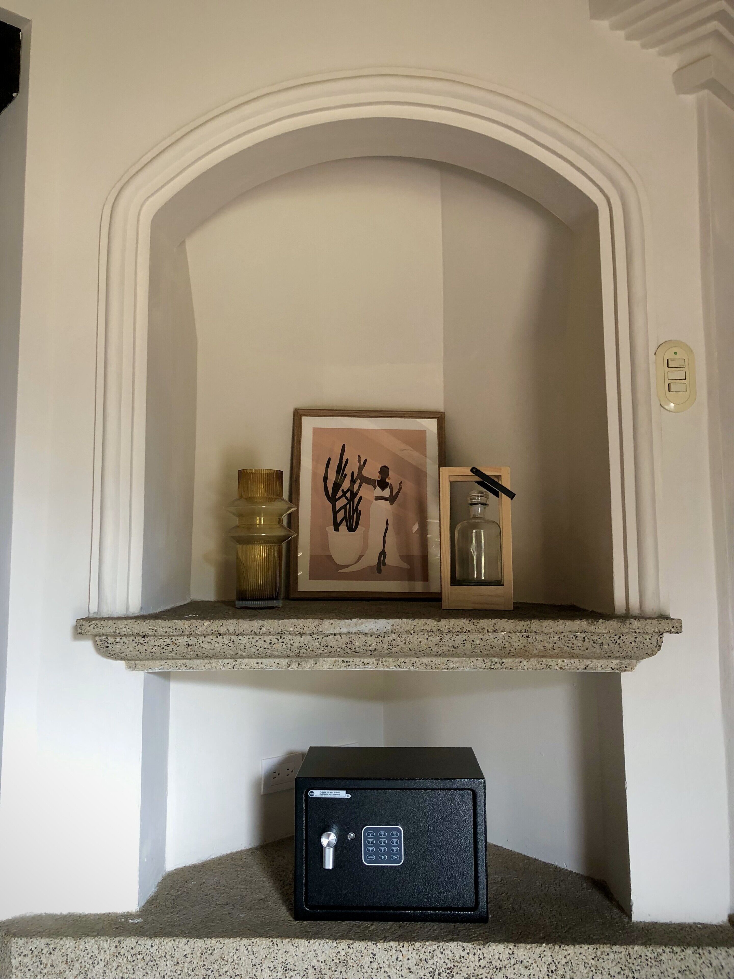 Une cheminée surmontée d’un tableau encadré, une petite étagère avec une bougie et un bocal en verre, et un coffre-fort noir placé sous l’étagère.