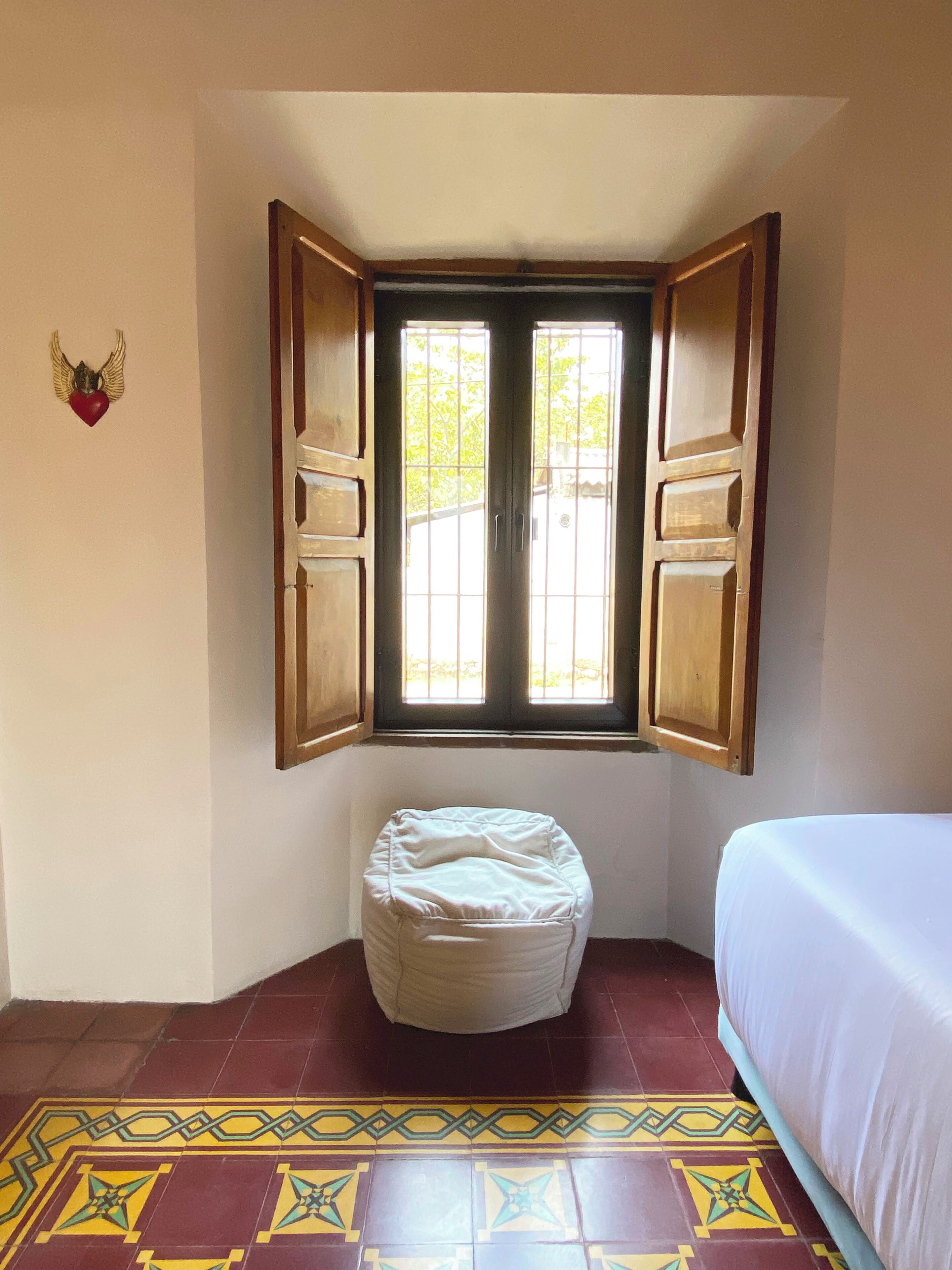 Une pièce avec une fenêtre, un lit, un petit tabouret et un élément décoratif mural.