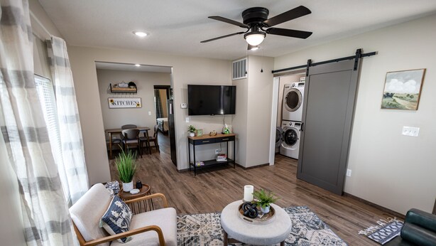 A modern living room with a sofa, armchair, and ottoman, a dining area, a kitchen with a TV, and a laundry area.