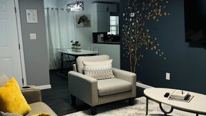 A modern living room with a grey sofa, a beige armchair, and a round coffee table. There is a dining area with a table and chairs, and a kitchen in the background.