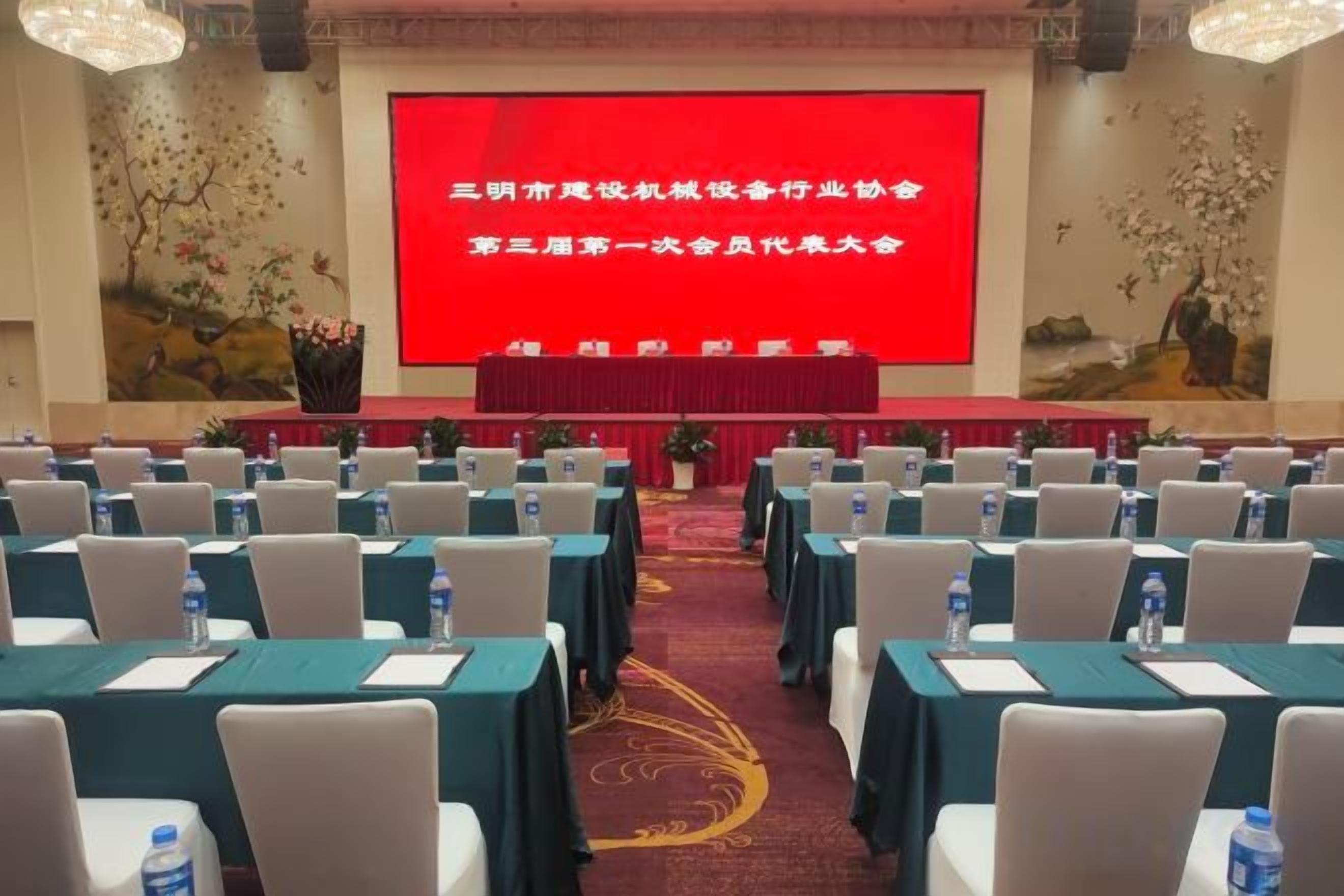 Un salón de conferencias con filas de mesas y sillas, una pantalla grande al frente y una alfombra roja.