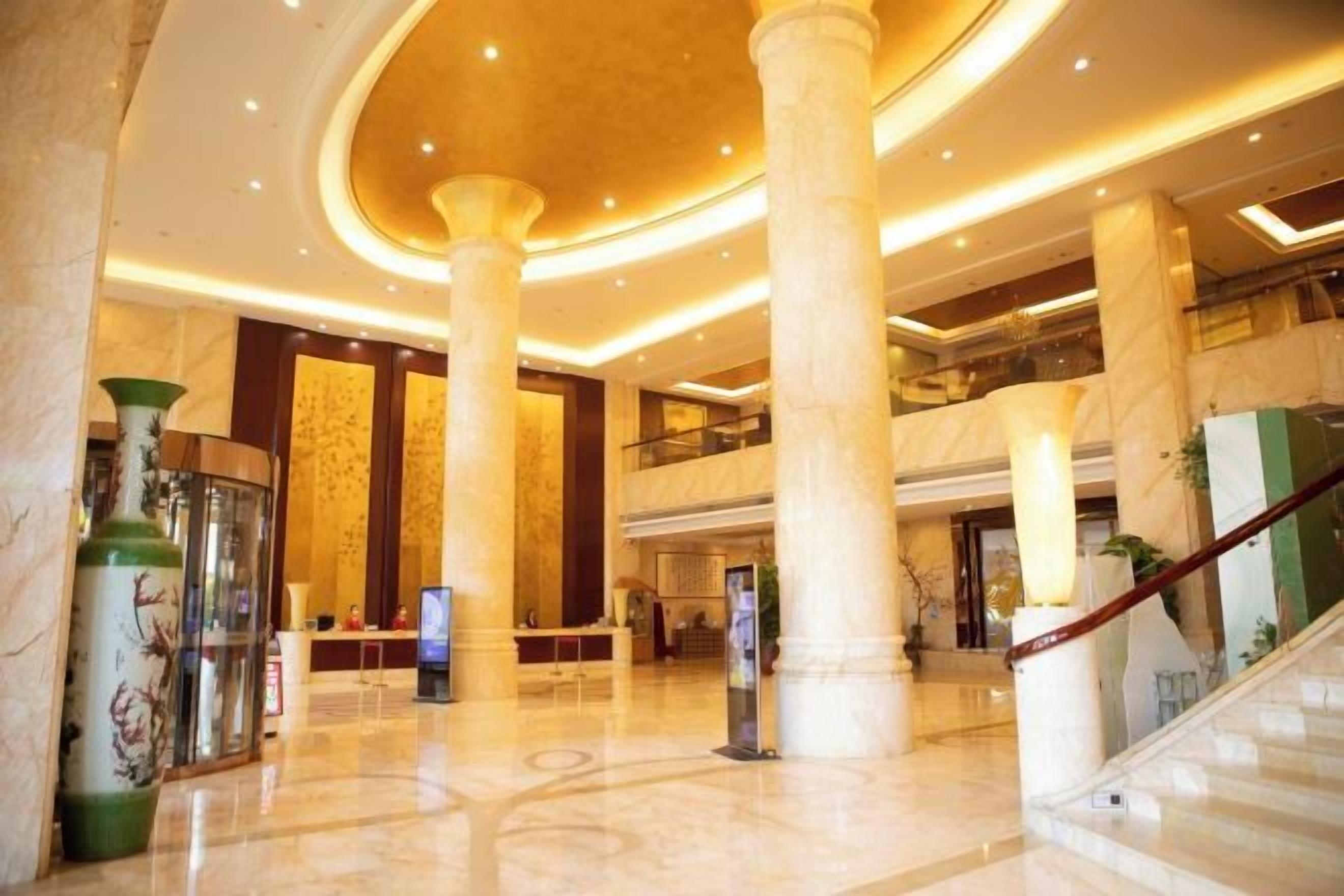 A spacious lobby with marble flooring, large columns, a staircase, and a high ceiling with recessed lighting.