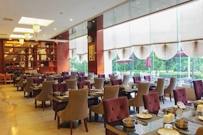 Un restaurant spacieux, doté de nombreuses tables et chaises, de grandes fenêtres avec des rideaux et d’un intérieur bien éclairé.