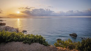 A coastal landscape with a sunset over the sea, rocky terrain, and sparse vegetation.