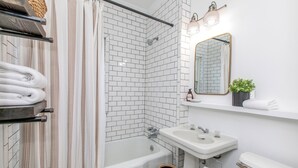 A modern bathroom with a corner shower, white tiled walls, a white sink, and a mirror with a plant on the shelf. - Modern Loft in Historic Birdtown - Unit 4 (Lakewood)