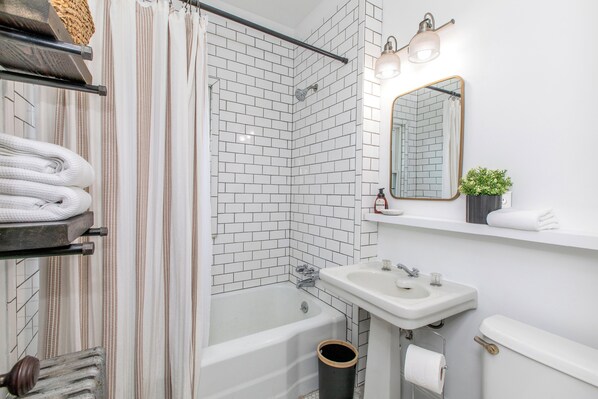 A modern bathroom with a corner shower, white tiled walls, a white sink, and a mirror with a plant on the shelf.