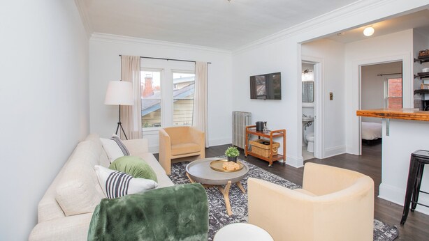 A living room with a sofa, chairs, a coffee table, and a view of the outside. There is a kitchen area with a counter and a hanging light fixture. - Modern Loft in Historic Birdtown - Unit 4 (Lakewood)