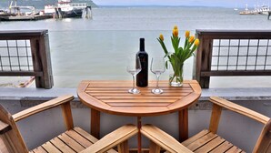 Een houten tafel met twee wijnglazen en een fles, omringd door stoelen, met uitzicht op een rustig wateroppervlak waar bootjes in de verte liggen.