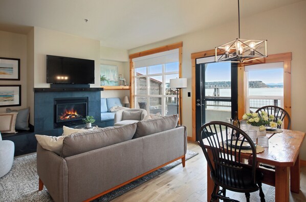 A living room with a fireplace, a television, a sofa, and a dining area with a view of a body of water.