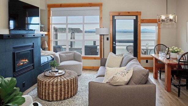 A cozy living room with a fireplace, comfortable seating, a dining area, and large windows overlooking a body of water. - Waterfront 1 bedroom condo - private deck offering spectacular views (Langley)