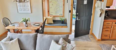 Un salon confortable avec un canapé, une table à manger et des chaises, une carte accrochée au mur et une porte donnant sur la cuisine.