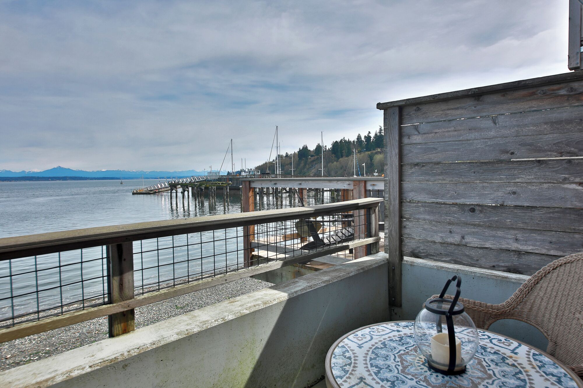 A balcony with a view of a dock and water, a wicker chair, and a table with a candle.