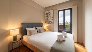 A neatly made bed with white linens and two pillows, a bedside lamp, and a window with a view of buildings and greenery. - Elegant Duplex Apt in Downtown by Lisbeyond (Porto)