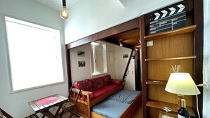 A compact room with a loft bed, a small table, and a chair. There is a ladder leading to the loft bed, and a shelf with a lamp and wine glasses. - Studio Edifício Leblon (Leblon Rio de Janeiro)