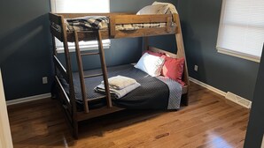 Une chambre avec un lit superposé, un ventilateur de plafond, un plancher en bois et deux fenêtres équipées de stores.