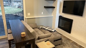 Un salon avec une table en bois foncé, un canapé d’angle gris, un plaid à carreaux, un téléviseur à écran plat fixé à un mur en briques et une cheminée.