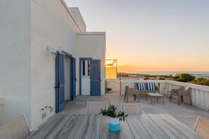 Une terrasse avec une table, des chaises et un pot de fleurs, d’où l’on peut admirer le coucher de soleil et la mer.
