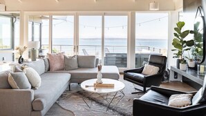 Een moderne woonkamer met een grijze hoekbank, een zwarte leren fauteuil en een ronde salontafel. Grote ramen bieden uitzicht op de zee.
