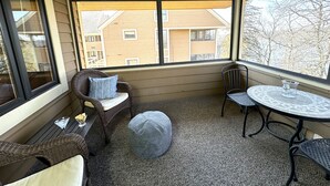 A covered patio with wicker furniture, a round table, and a view of a building through large windows.