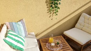 Een zonnig terras met twee zithoeken met kussens, een houten tafel met drankjes en een potplant.