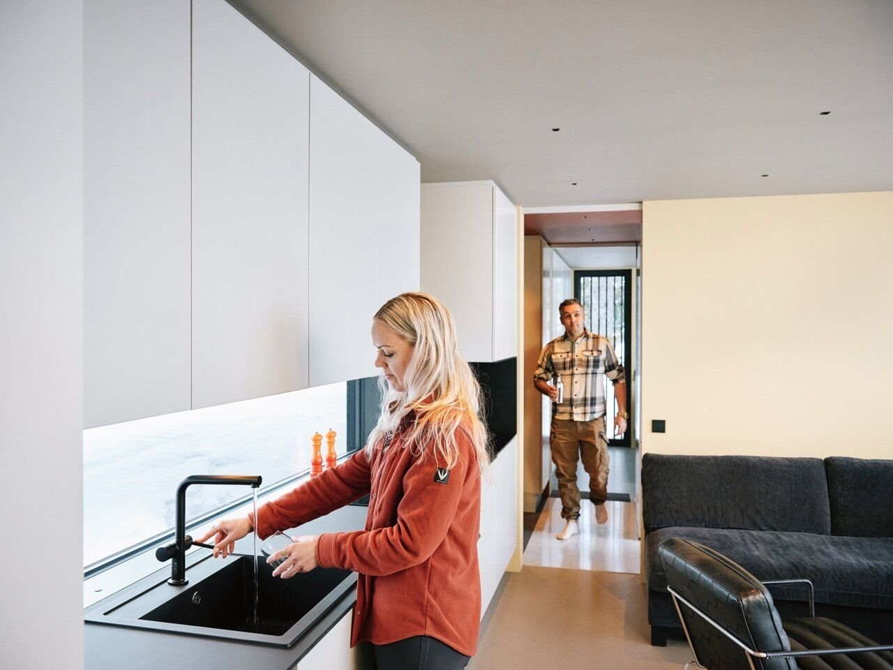 A modern kitchen with a woman washing dishes and a man walking in the background.