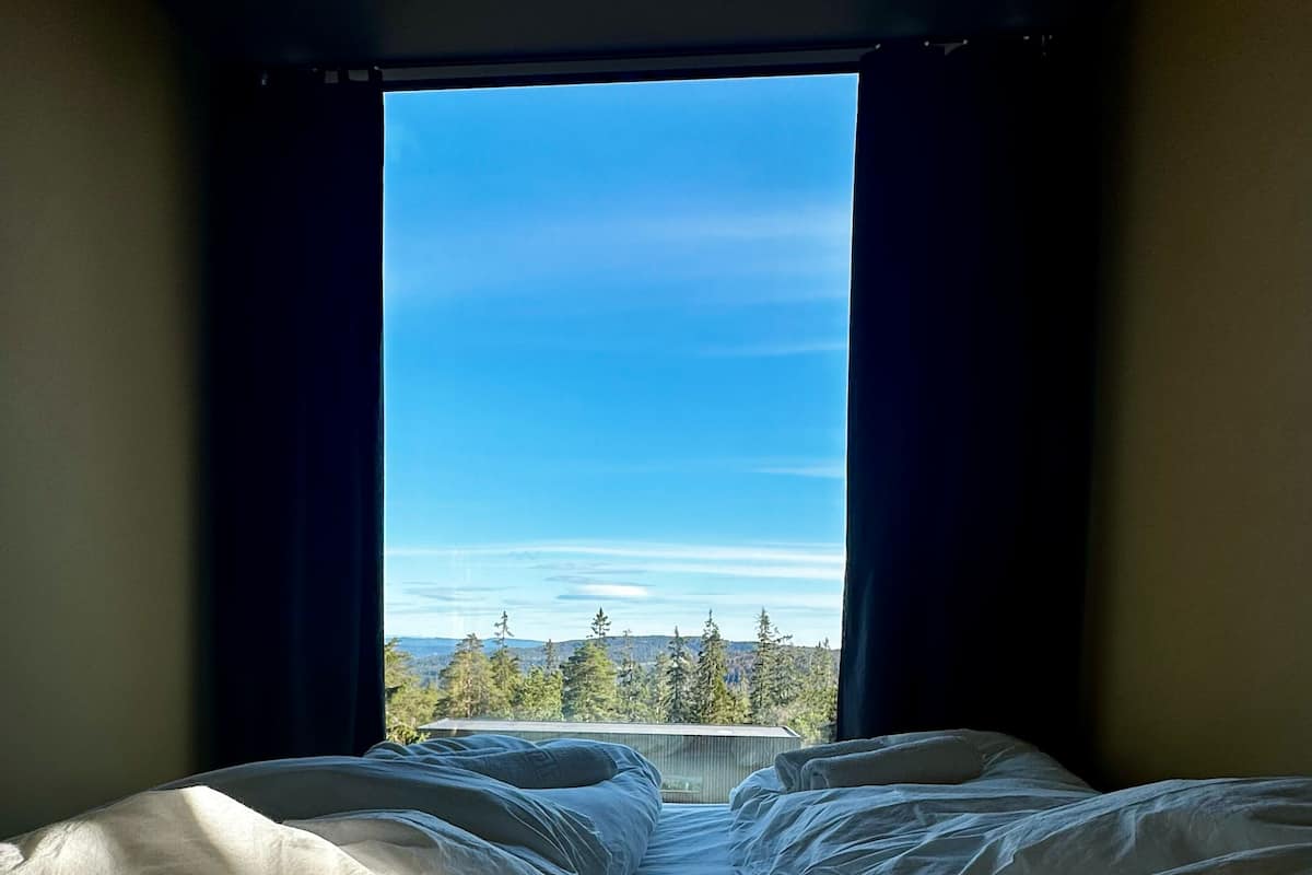 Una habitación con una cama orientada a un ventanal, cortinas y vistas de árboles y montañas.