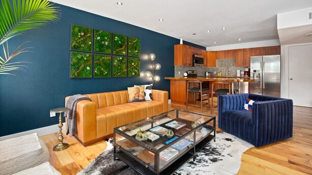 Una sala de estar moderna con un sofá, una mesa de centro y un sillón azul. Se observa una cocina al fondo con armarios de madera y un refrigerador.