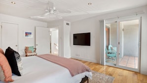 Un dormitorio con cama, televisor, ventilador de techo, puerta corrediza de vidrio a un balcón y una silla.