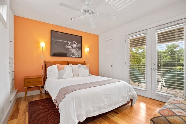 A bedroom with a bed, a nightstand, a ceiling fan, a large window with blinds, and a view of greenery outside.