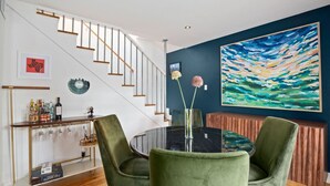 Un comedor con mesa de vidrio, sillas tapizadas en verde, una consola de madera con botellas y copas, un cuadro abstracto grande en la pared y una escalera con barandillas de madera.