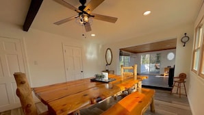 A spacious dining area with a large wooden table and benches, a ceiling fan, and a view into another room.