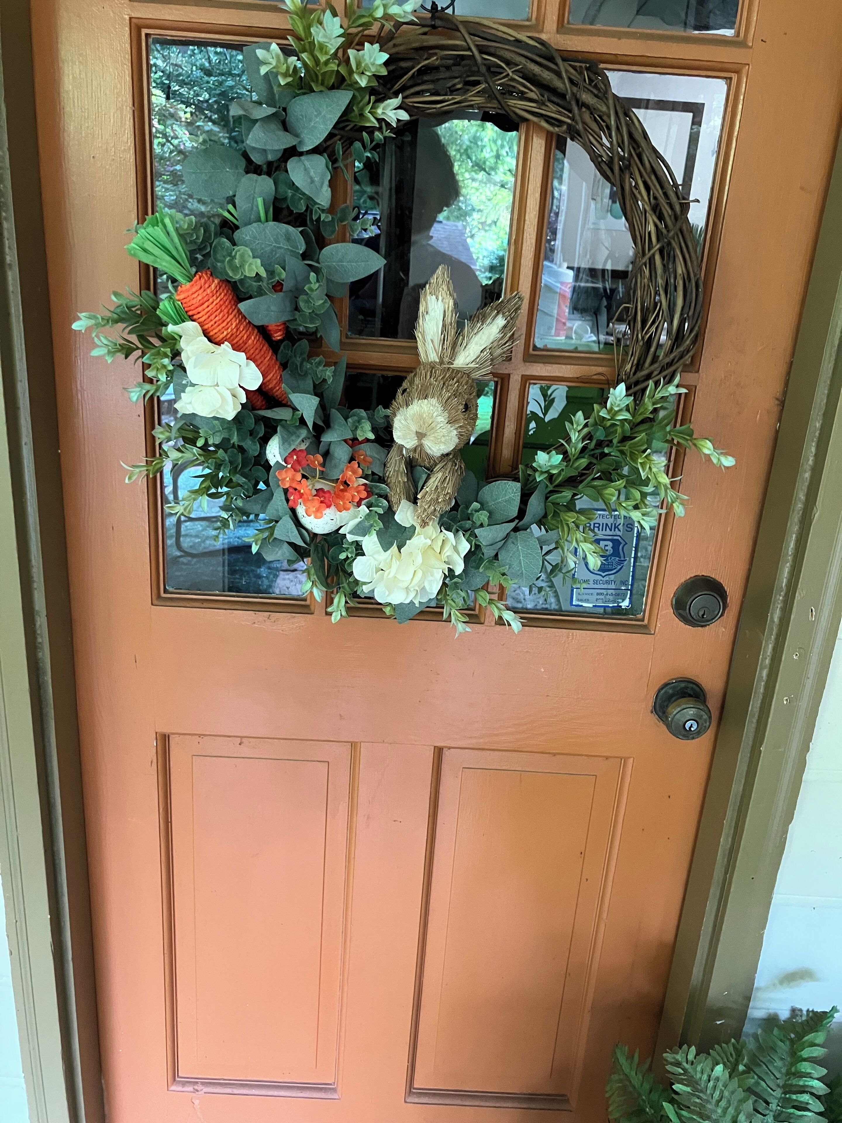 Une porte en bois avec une fenêtre, un carillon et une guirlande décorée de fleurs artificielles et d’un petit lapin.