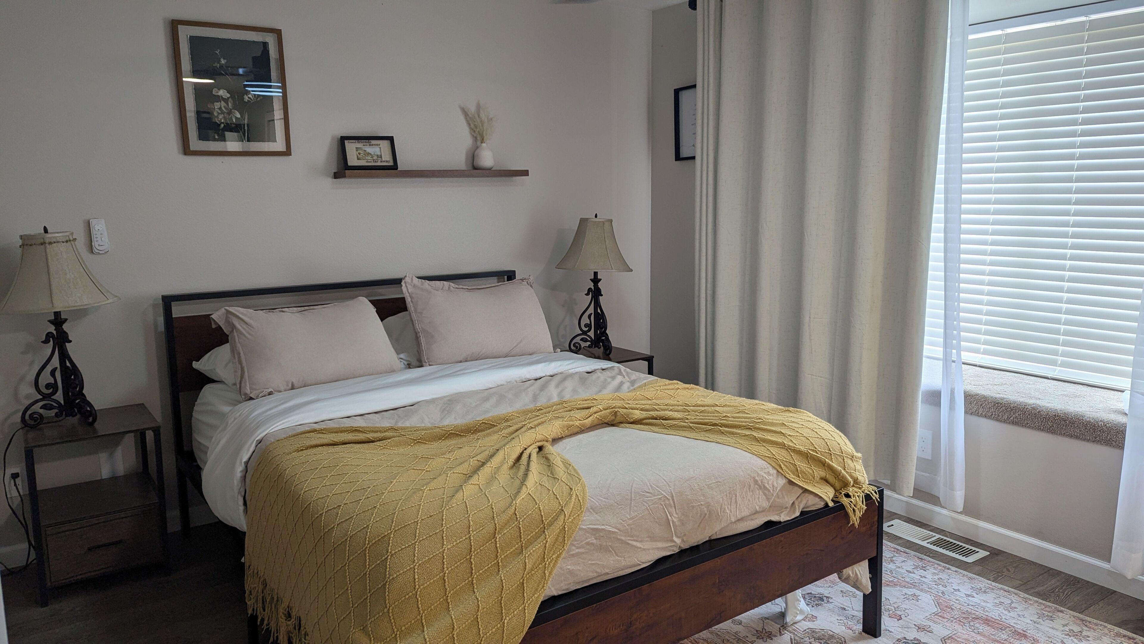 Una cama bien hecha con un edredón amarillo, dos lámparas de noche, un pequeño estante con adornos y una ventana con persianas.