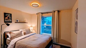 A bedroom with a bed, a bedside lamp, a window with curtains, and wall art.