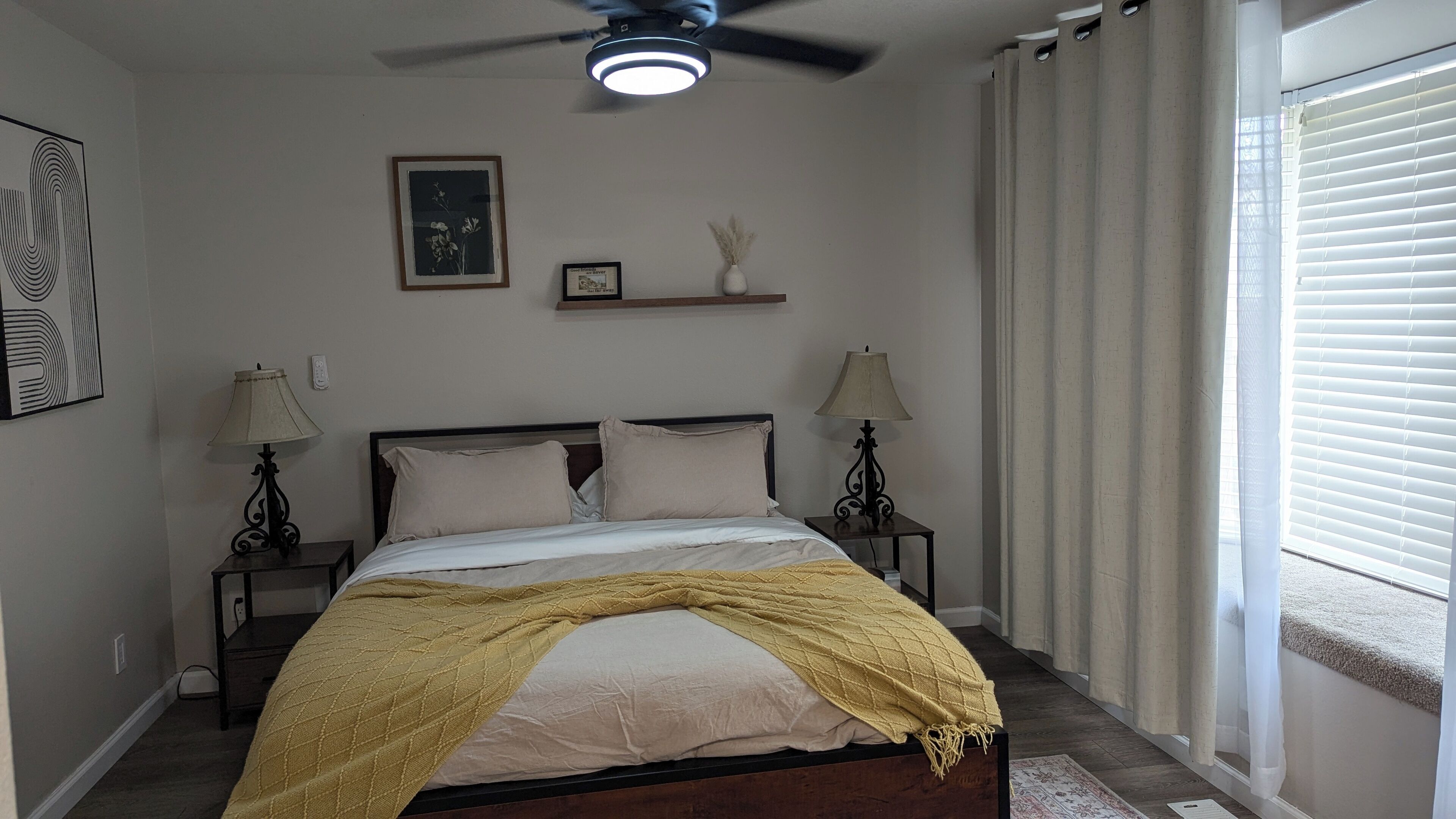 Un dormitorio con cama, dos mesitas de noche con lámparas, un ventilador de techo, una ventana con persianas y un estante montado en la pared con objetos decorativos.