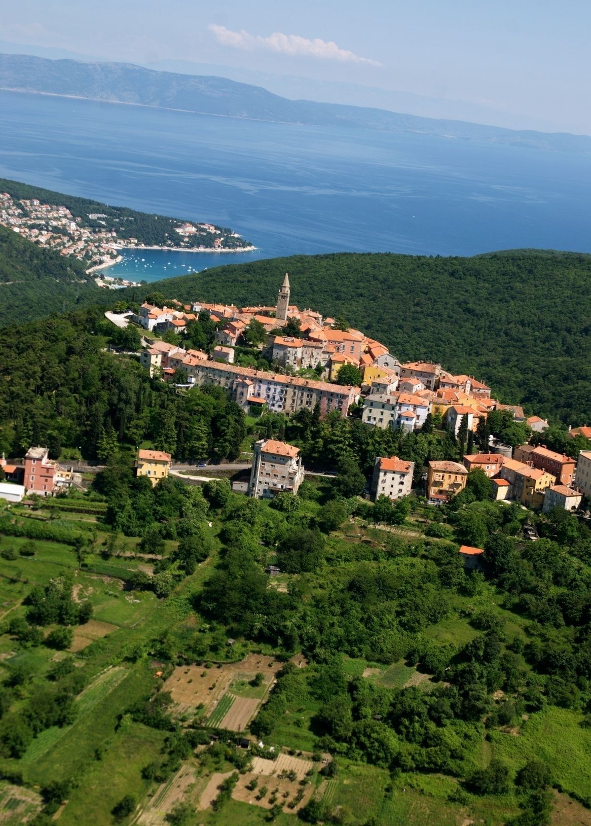 An aerial view of a coastal town with buildings clustered on a hillside, overlooking a bay with boats.