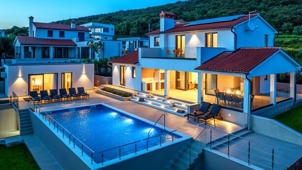 A modern villa with a swimming pool, outdoor seating, and a view of a lush hillside.