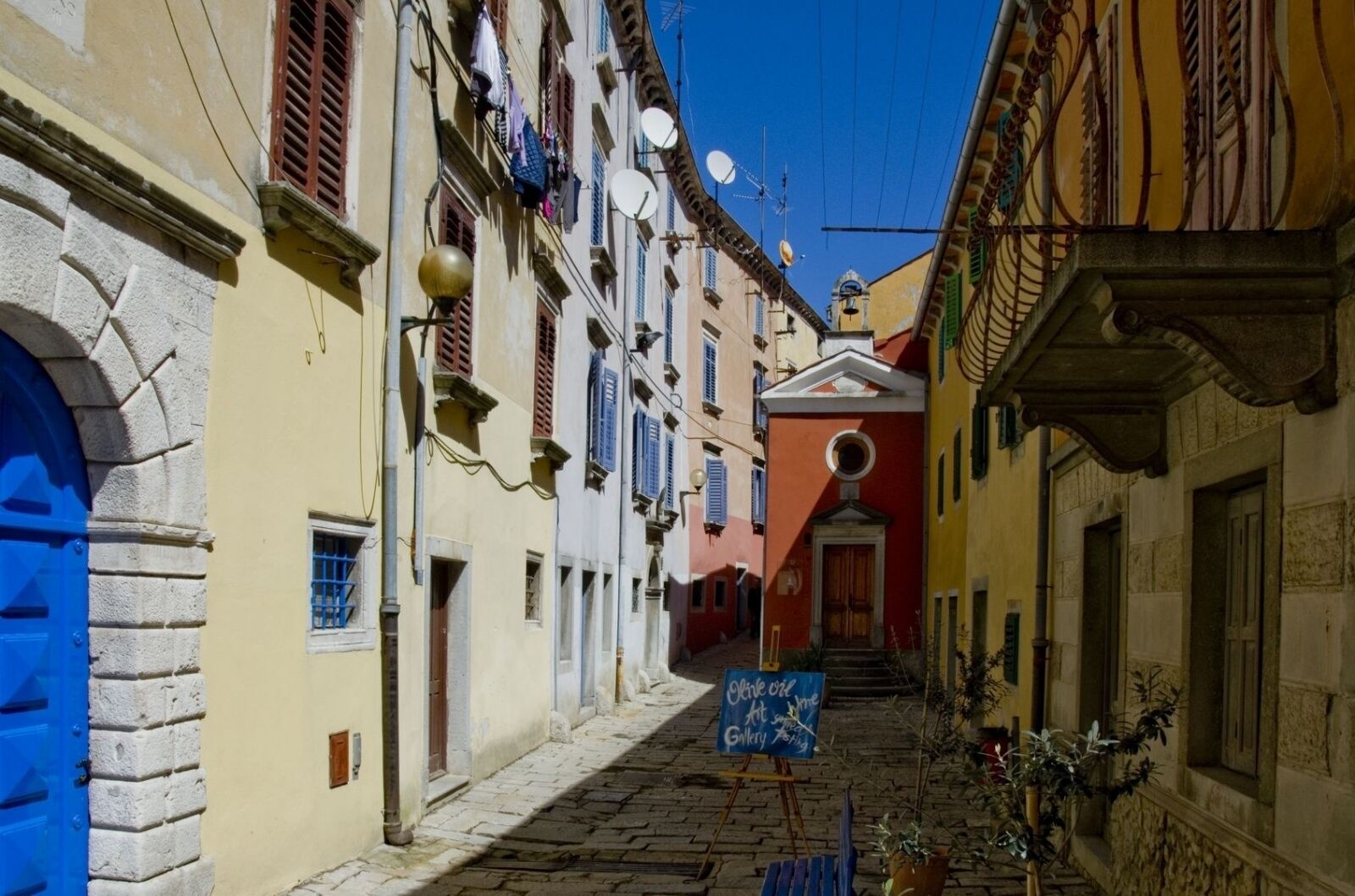 A narrow cobblestone street lined with colorful buildings, a red church, and a signboard.