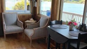 A cozy cabin with wooden walls and floors, a dining table with chairs, and two armchairs. Large windows offer views of a lake and greenery.