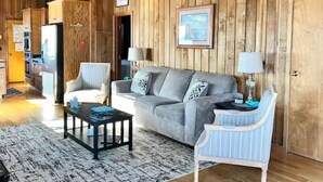 A cozy living room with a grey sofa, a white armchair, and a coffee table. Wooden paneling adorns the walls, and a framed picture hangs above the sofa.