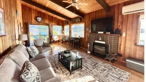A cozy cabin interior with a fireplace, wooden walls, a ceiling fan, and a large window overlooking a scenic view.