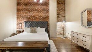 A bedroom with a brick wall, a tufted headboard, a wooden bed frame, a bedside table, a floor lamp, and a dresser with drawers.
