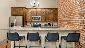 A kitchen with wooden cabinets, a granite countertop, a stainless steel refrigerator, a microwave, a stove, and a chandelier. There are three bar stools.