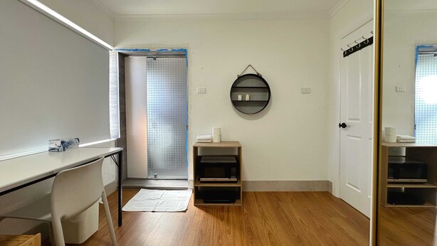 Une pièce avec un plancher en bois, un bureau blanc, une chaise, une petite étagère contenant des articles de toilette, une étagère ronde fixée au mur et une porte dotée d’un miroir.
