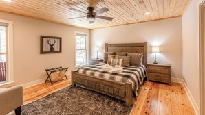 A bedroom with a wooden ceiling, a bed with a checkered bedspread, two bedside tables with lamps, and a framed deer head on the wall.