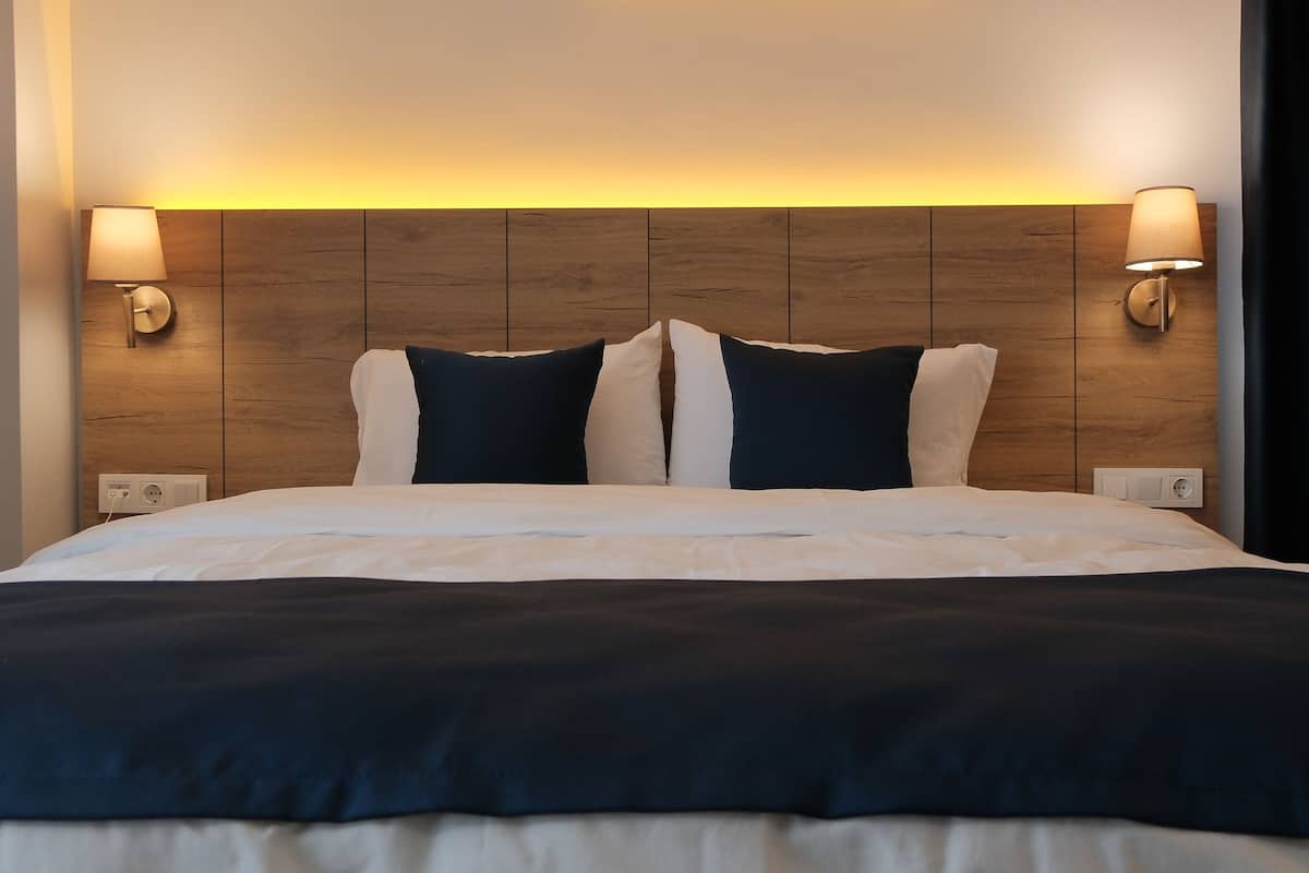 A hotel room with a wooden headboard, two bedside lamps, and a bed with white bedding and dark pillows.