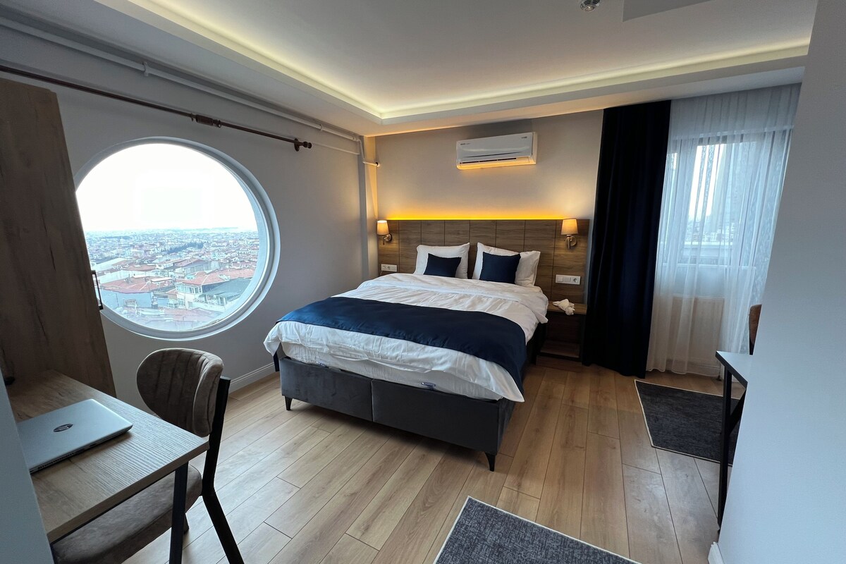 Une chambre d’hôtel avec un grand lit, un bureau, une chaise, une fenêtre ronde et un appareil de climatisation.