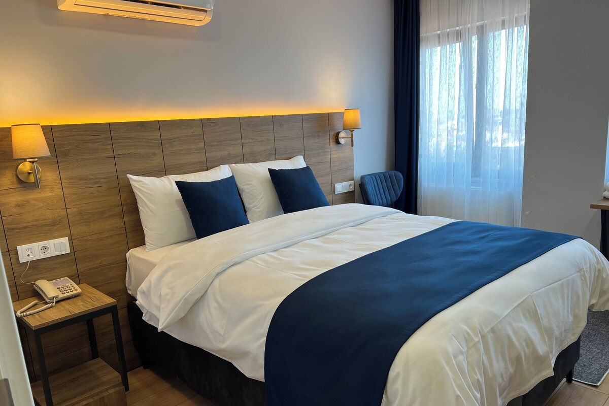 Habitación de hotel con cama, lámpara de noche, teléfono y ventana con cortinas.