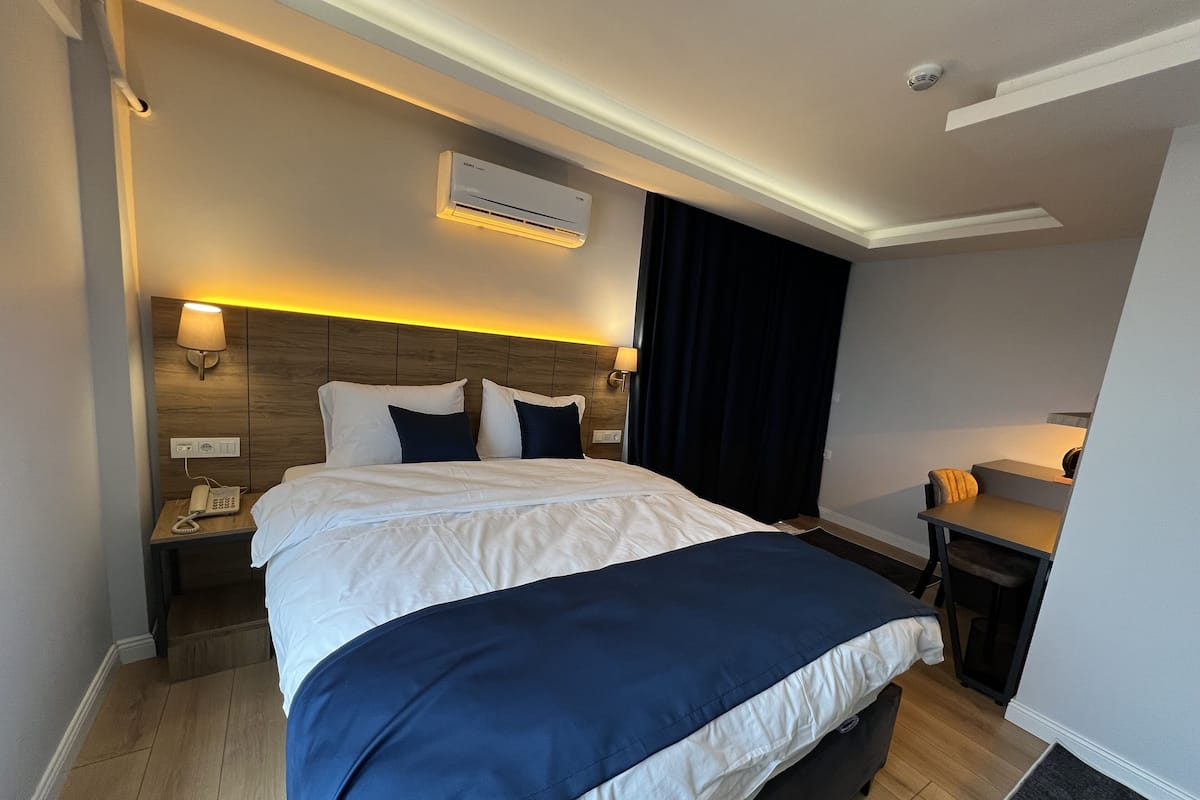 A hotel room with a large bed, bedside tables, an air conditioning unit, a desk with a chair, and a telephone.