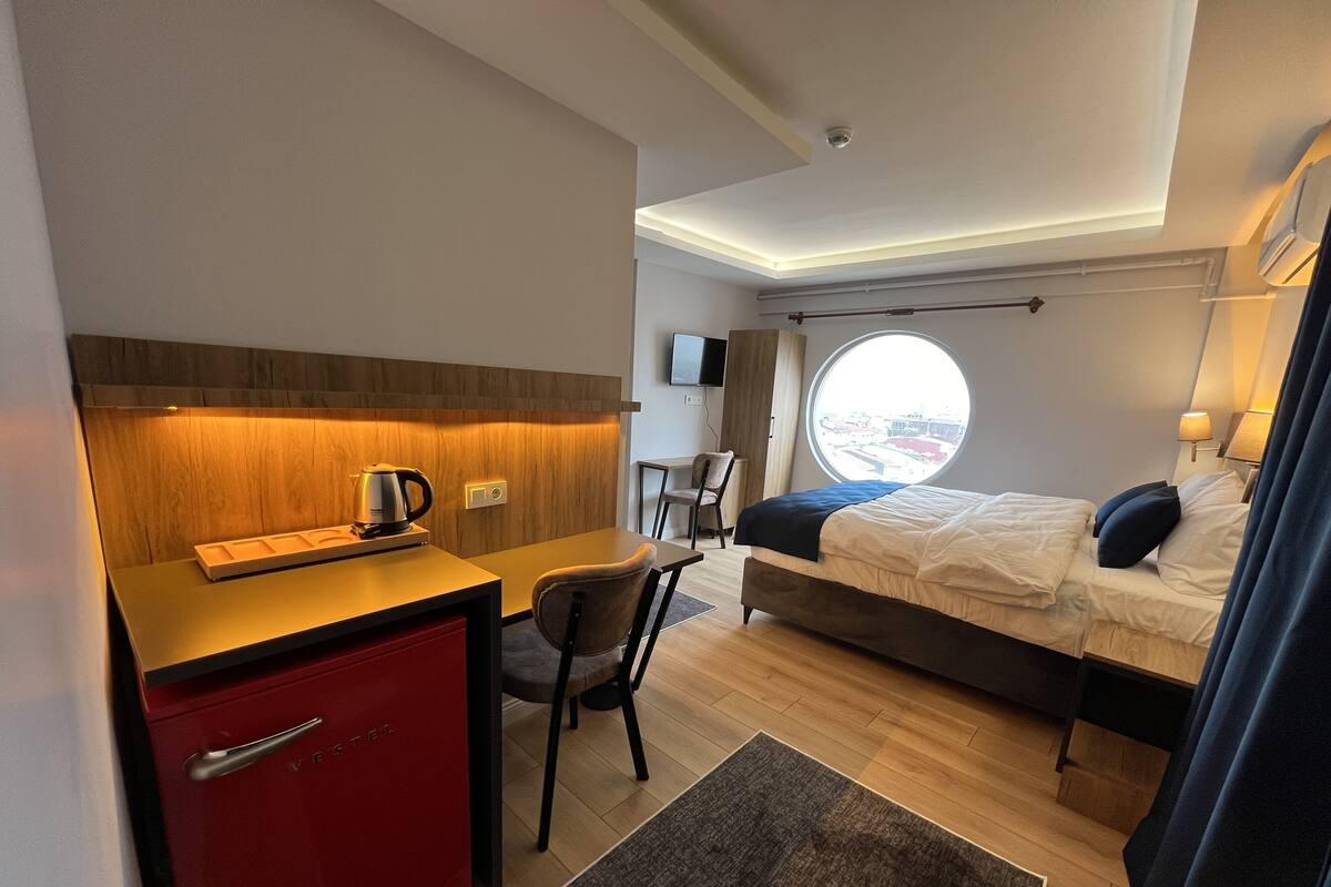 A compact hotel room with a bed, a desk with a kettle, a chair, a red minibar, and a window with a view of the outside.